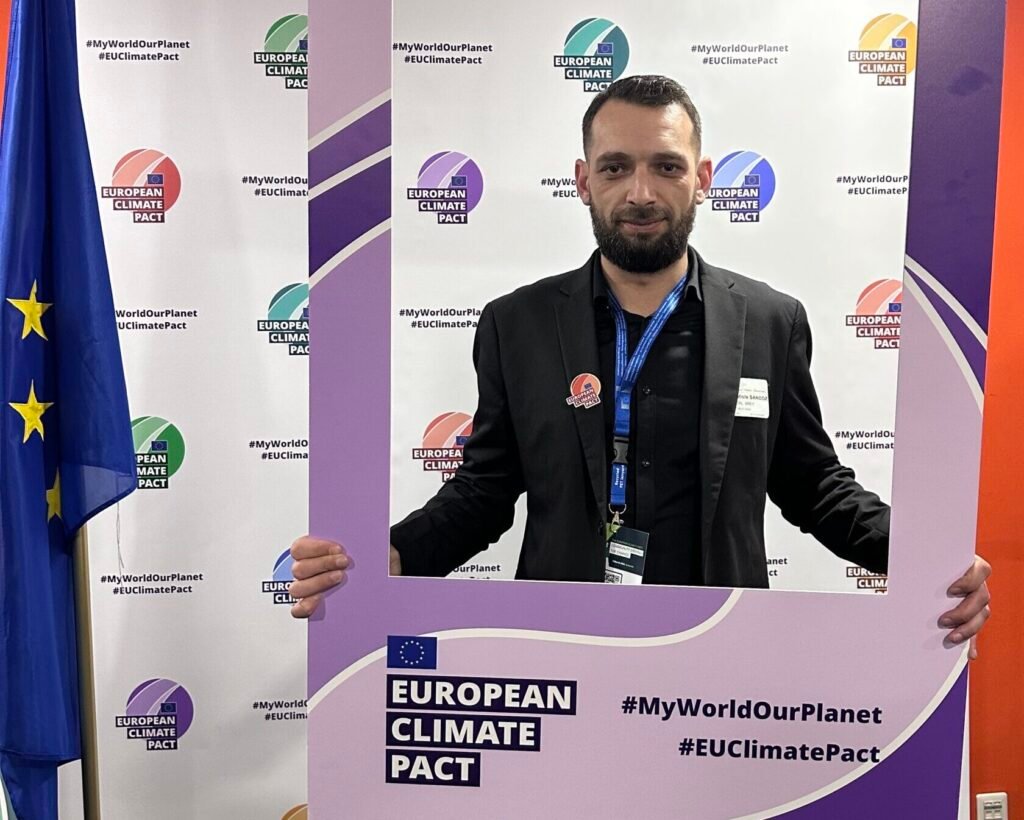 Jean-Baptiste Sandoz tenant un cadre de l’European Climate Pact avec les hashtags #MyWorldOurPlanet et #EUClimatePact, devant un fond avec le drapeau de l’UE et les logos officiels.